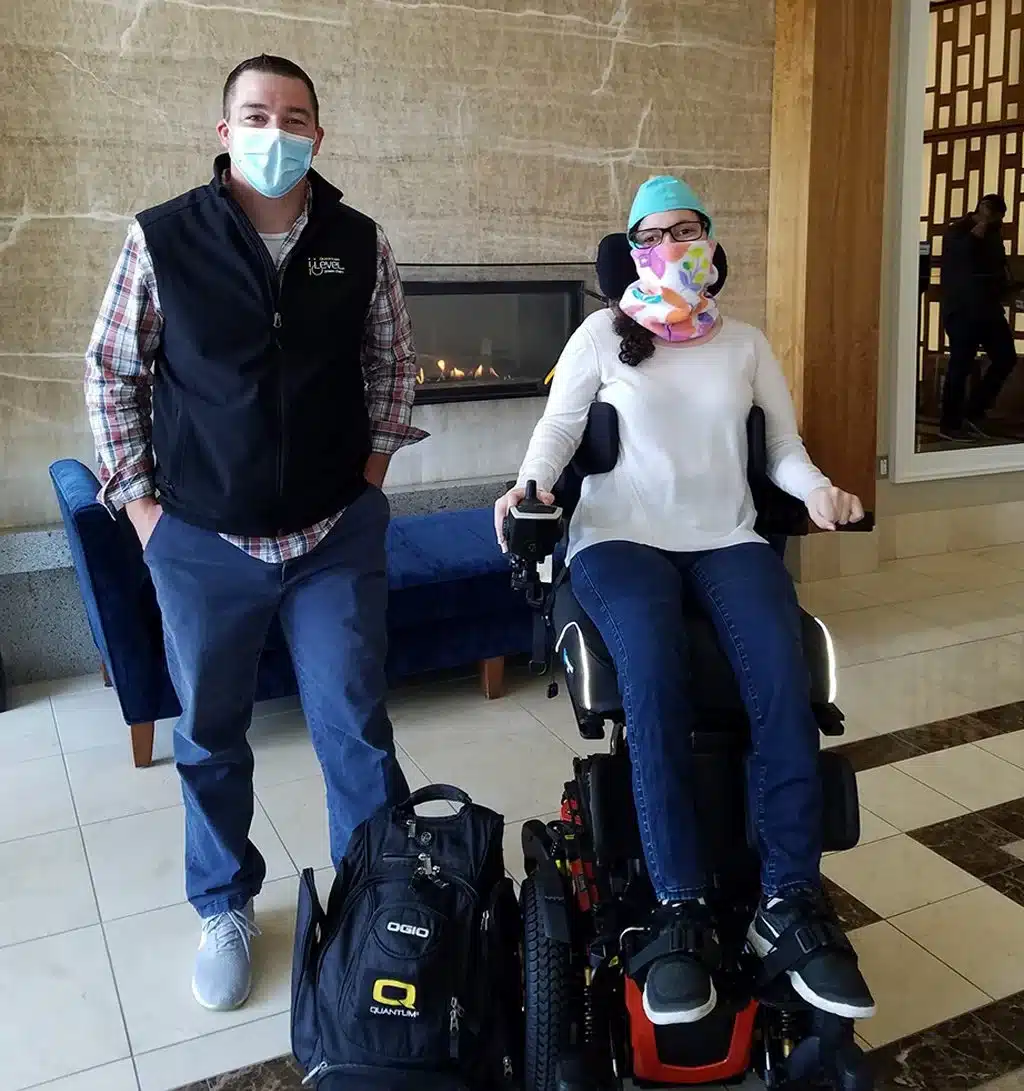 ilevel2 A man in a vest stands beside a woman in an electric wheelchair, with a modern fireplace in the background and a couch nearby.